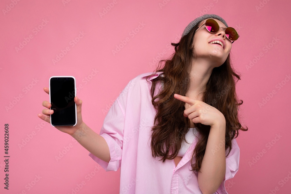 attractive young brunette woman wearing stylish pink shirt grey hat and colourful sunglasses isolated over pink background holding in hand and showing mobile phone with empty display for mockup attractive young brunette woman wearing stylish pink shirt grey hat and colourful sunglasses isolated over pink background holding in hand and showing mobile phone with empty display for mockup