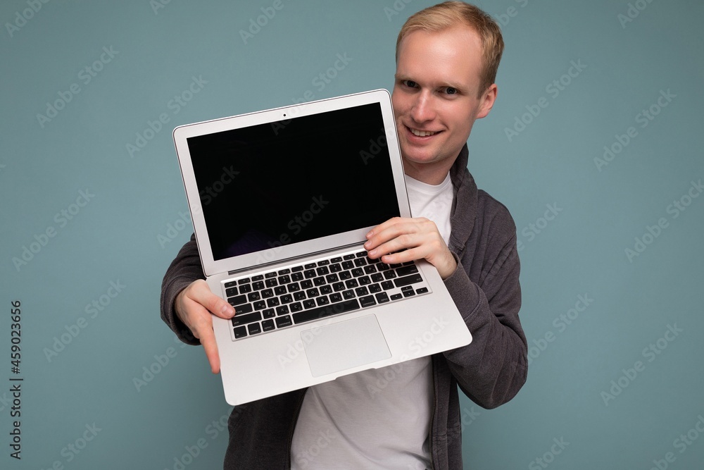 Close-up portrait of handsome smiling blonde man holding computer laptop with empty monitor screen with mock up and copy space wearing casual clothes looking at camera isolated over blue background Close-up portrait of handsome smiling blonde man holding computer laptop with empty monitor screen with mock up and copy space wearing casual clothes looking at camera isolated over blue background