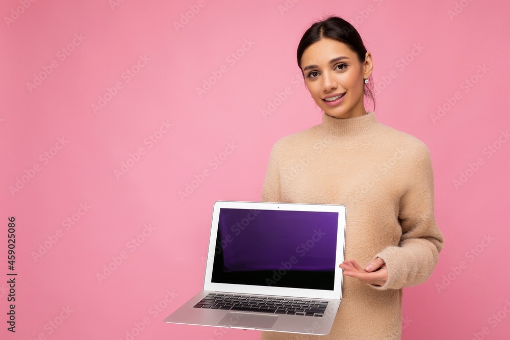 beautiful young woman with dark hair looking at camera holding computer laptop with empty monitor screen with mock up and copy space wearing light colour sweater isolated on pink wall background beautiful young woman with dark hair looking at camera holding computer laptop with empty monitor screen with mock up and copy space wearing light colour sweater isolated on pink wall background