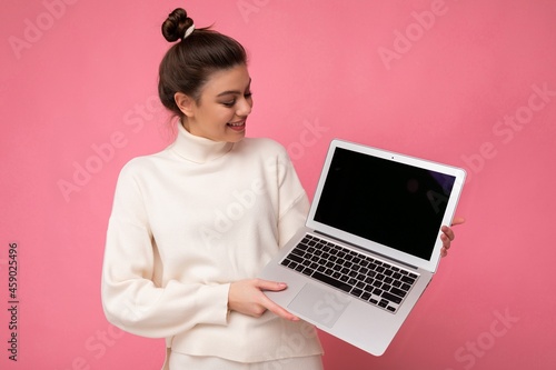 Photo of beautiful charming smiling woman with gathered brunette hair wearing white sweater holding computer laptop and looking at the netbook isolated over pink wall background photo