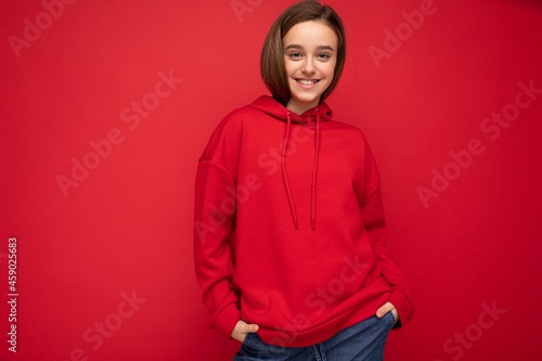 Photo shot of pretty cool happy smiling brunette little girl wearing trendy red hoodie standing isolated over red background wall looking at camera © Ivan Traimak