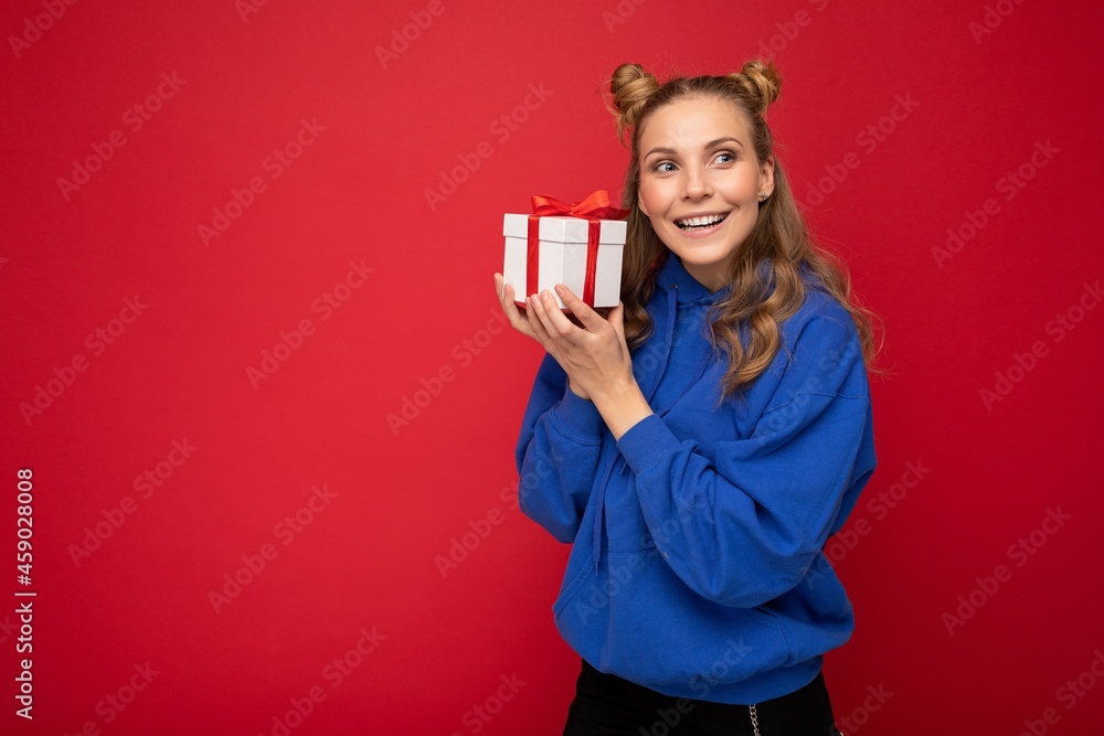 Shot of attractive happy smiling young blonde woman isolated over red background wall wearing blue trendy hoodie holding gift box and looking to the side Shot of attractive happy smiling young blonde woman isolated over red background wall wearing blue trendy hoodie holding gift box and looking to the side