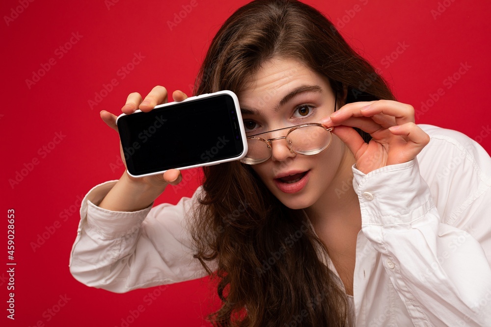 beautiful young brunette woman wearing white shirt and optical glasses isolated over red background holding in hand and showing mobile phone with empty screen for cutout looking at camera beautiful young brunette woman wearing white shirt and optical glasses isolated over red background holding in hand and showing mobile phone with empty screen for cutout looking at camera