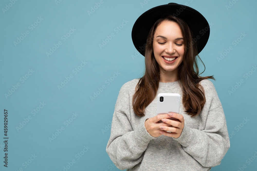 Attractive young smiling woman wearing black hat and grey sweater holding smartphone looking down isolated on background Attractive young smiling woman wearing black hat and grey sweater holding smartphone looking down isolated on background