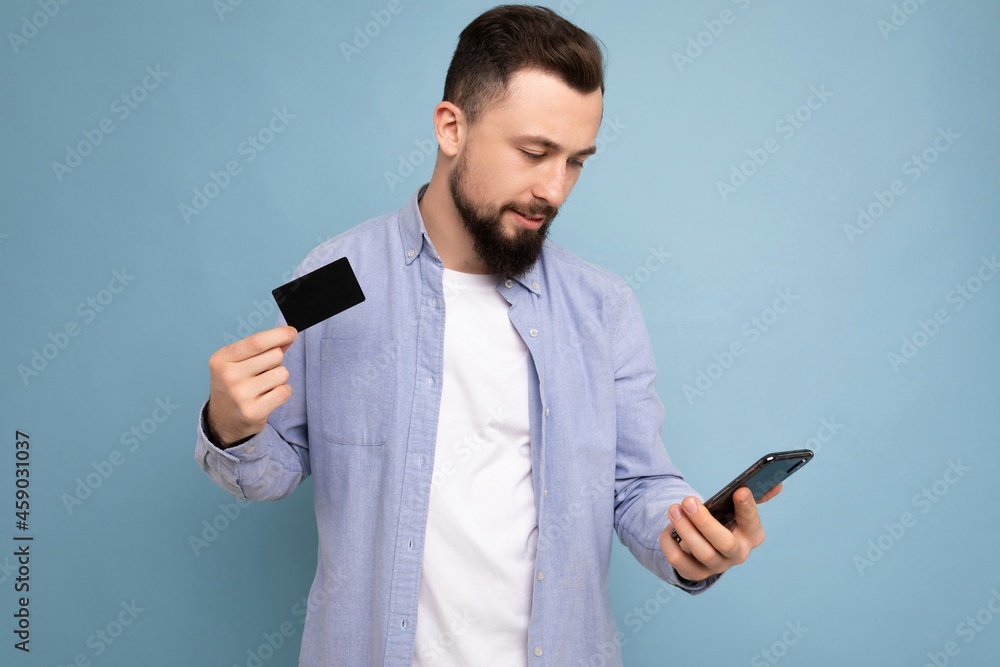 closeup Photo of good looking attractive smiling brunet unshaven young man wearing casual blue shirt and white t-shirt isolated over blue background wall holding credit card and using mobile phone closeup Photo of good looking attractive smiling brunet unshaven young man wearing casual blue shirt and white t-shirt isolated over blue background wall holding credit card and using mobile phone