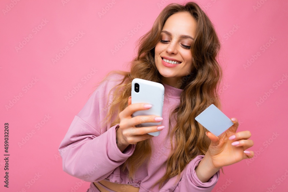 Beautiful smiling positive young blonde curly woman wearing pink clothes isolated over pink background using mobile phone paying online shopping through credit card looking at smartphone display Beautiful smiling positive young blonde curly woman wearing pink clothes isolated over pink background using mobile phone paying online shopping through credit card looking at smartphone display
