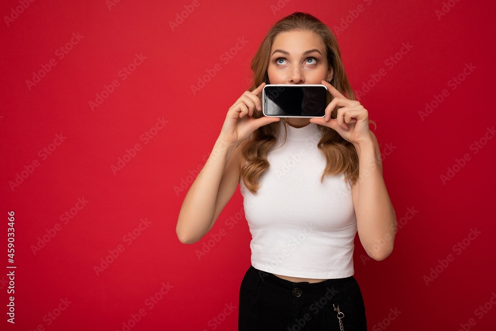 attractive young blonde female person wearing white t-shirt isolated on red background with copy space holding smartphone showing phone in hand with empty screen for cutout looking up attractive young blonde female person wearing white t-shirt isolated on red background with copy space holding smartphone showing phone in hand with empty screen for cutout looking up