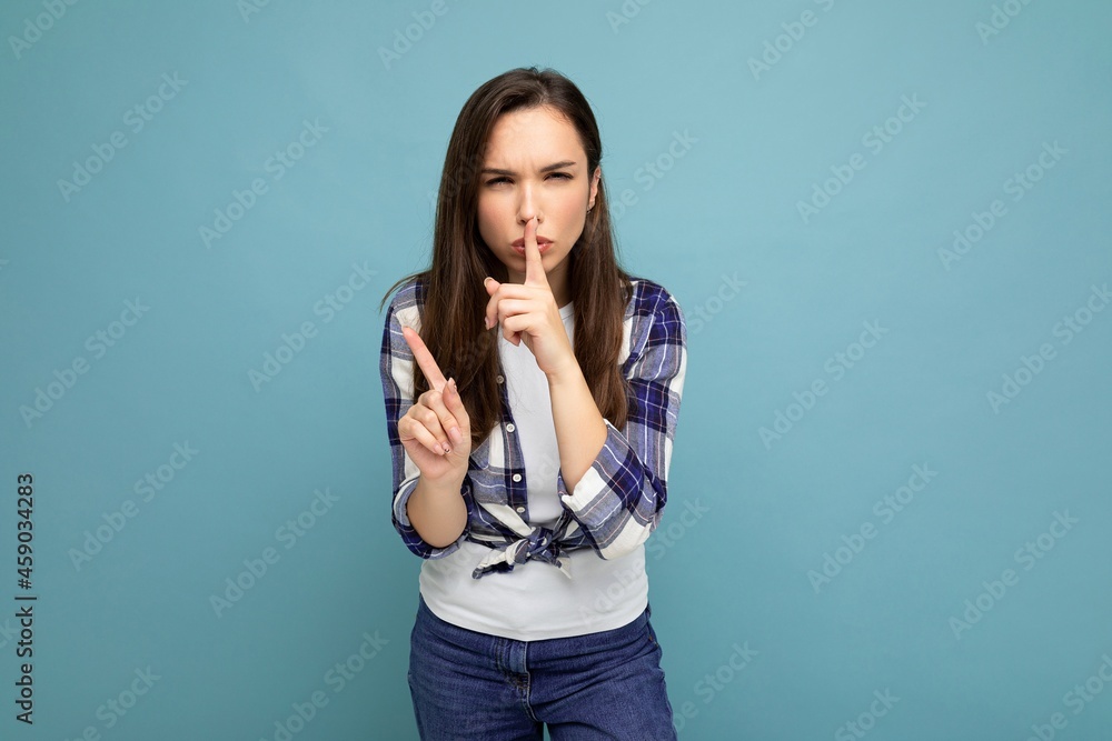 Young serious angry beautiful brunet woman with sincere emotions wearing trendy check shirt standing isolated on blue background with empty space and showing shhh gesture. Keep secret concept Young serious angry beautiful brunet woman with sincere emotions wearing trendy check shirt standing isolated on blue background with empty space and showing shhh gesture. Keep secret concept