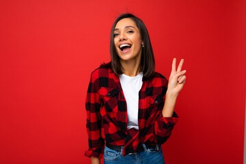 Young happy positive cool beautiful brunette woman with sincere emotions wearing white t-shirt and stylish red check shirt isolated over red background with copy space and showing two fingers