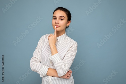 Portrait of young attractive beautiful thoughtful brunette woman with sincere emotions wearing casual srtriped white shirt isolated over blue background with copy space and thinking. Idea concept