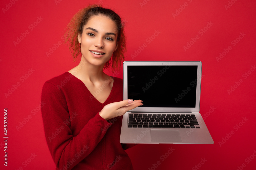 Close-up portrait of smiling happy beautiful dark blond woman holding laptop computer looking at camera showing at netbook keyboard wearing red sweater isolated over red wall background Close-up portrait of smiling happy beautiful dark blond woman holding laptop computer looking at camera showing at netbook keyboard wearing red sweater isolated over red wall background