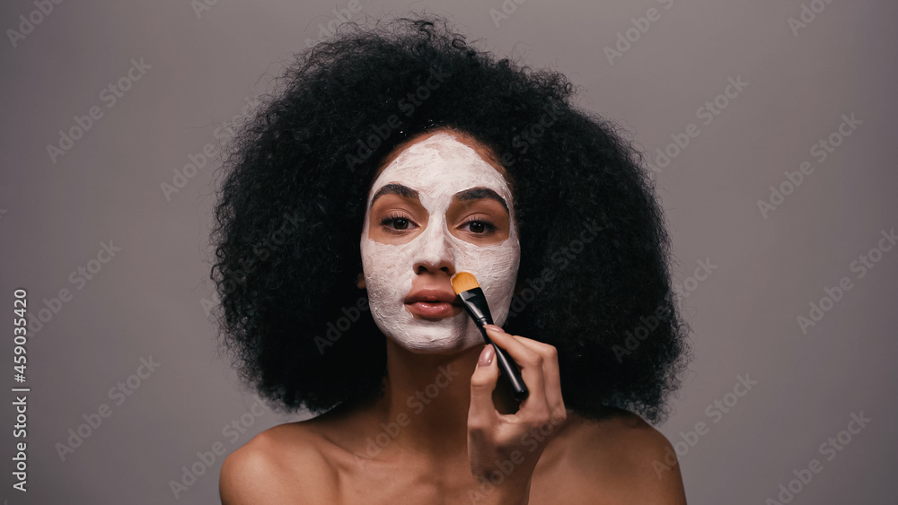 Obraz premium curly african american woman with bare shoulders applying clay mask isolated on grey