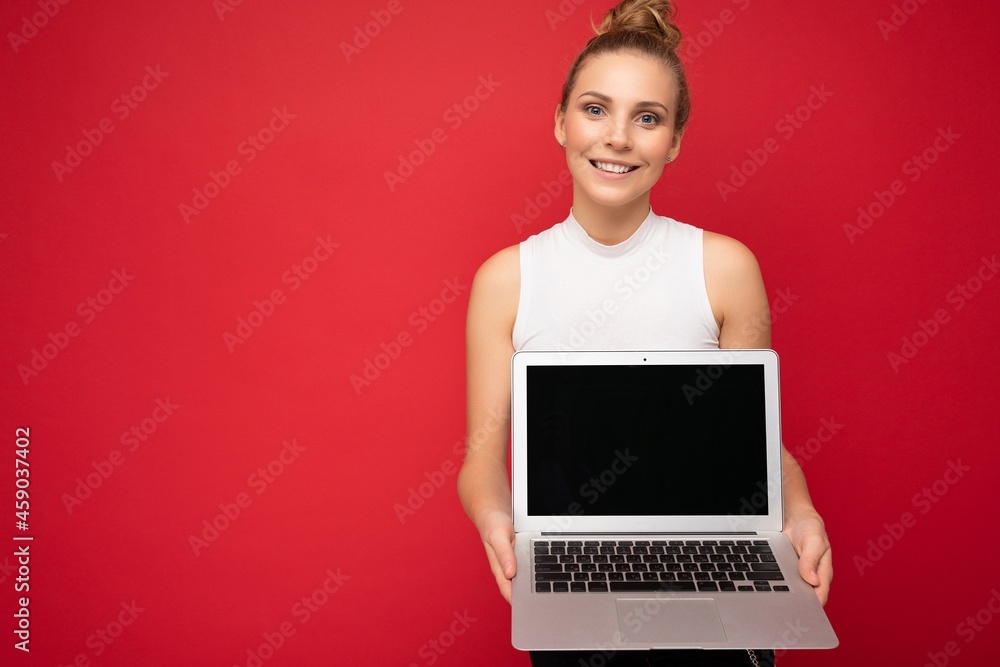 Smiling beautiful blond young woman with gathered hair looking at camera holding computer laptop with empty monitor screen with mock up and copy space wearing white t-shirt isolated over red wall Smiling beautiful blond young woman with gathered hair looking at camera holding computer laptop with empty monitor screen with mock up and copy space wearing white t-shirt isolated over red wall