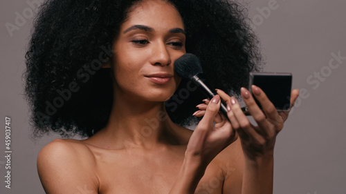 young african american woman applying face powder with cosmetic brush isolated on grey