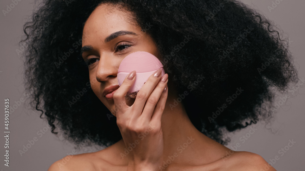 Naklejka premium curly african american woman using cleansing gadget on face isolated on grey