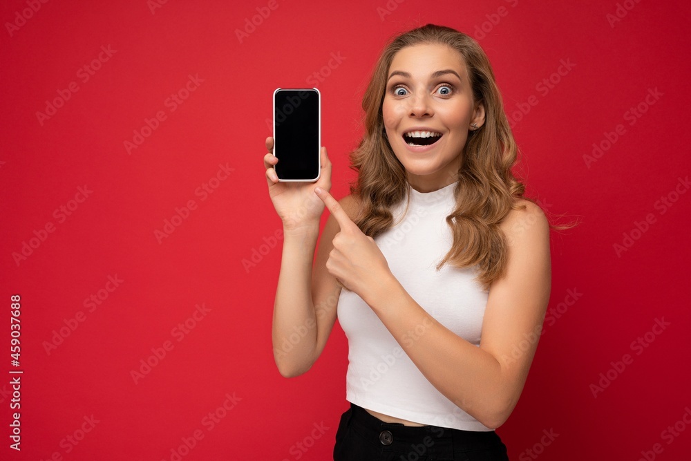 Shocked beautiful young blonde woman with open mouth wearing white t-shirt isolated on red background with copy space holding smartphone showing phone in hand with empty screen for mockup looking at Shocked beautiful young blonde woman with open mouth wearing white t-shirt isolated on red background with copy space holding smartphone showing phone in hand with empty screen for mockup looking at