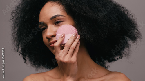 Fotografía curly african american woman using cleansing gadget on face isolated on grey