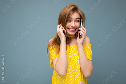 Closeup photo of pretty positive young blonde woman wearing stylish yellow summer dress standing isolated over blue background holding and talking on mobile phone with sincere emotions looking to the