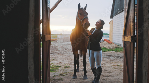 a young woman stands upright next to a horse she is holding by the halter in front of the entrance to the barn illuminated by natural light. High-quality photo photo