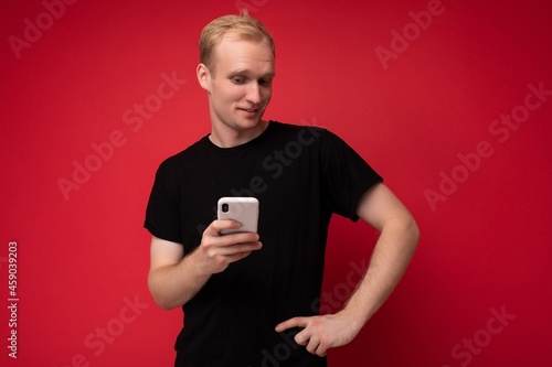 Photo of handsome smiling positive young blonde man isolated over red background wall wearing black t-shirt holding and using mobile phone writing sms looking at camera