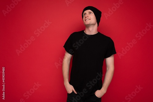 Handsome positive self-confident young man standing isolated over red background wall wearing casual black t-shirt for mockup and black hat and looking to the side