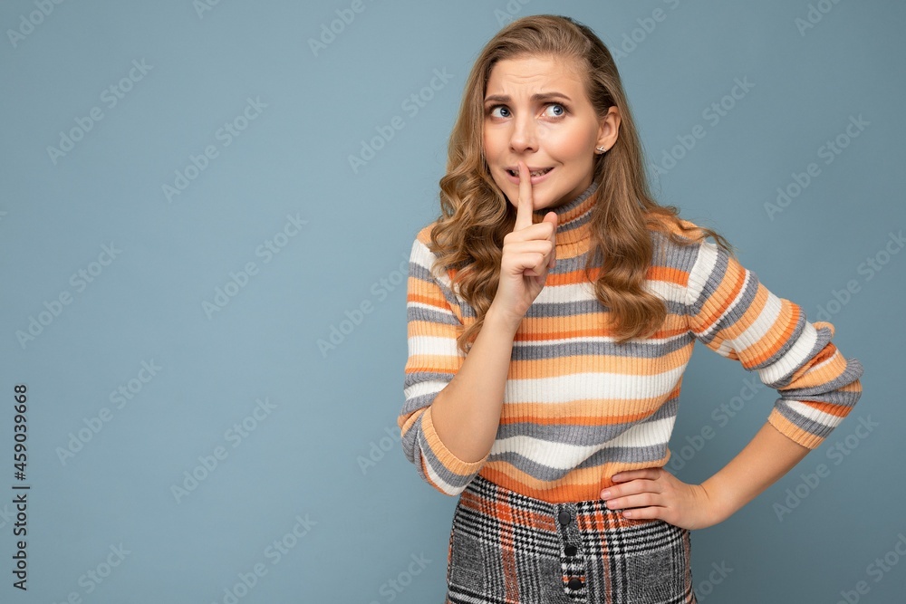 Portrait of young blonde beautiful curly woman with sincere emotions wearing striped sweater isolated on blue background with empty space and showing silence gesture with finger over lips Portrait of young blonde beautiful curly woman with sincere emotions wearing striped sweater isolated on blue background with empty space and showing silence gesture with finger over lips