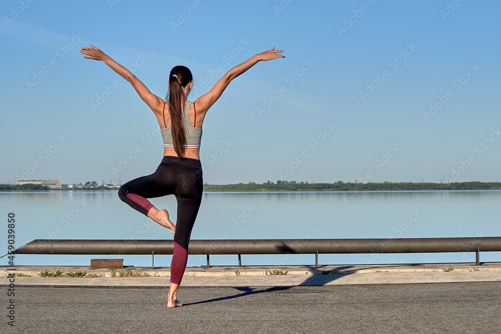 Fototapeta premium A young woman does a warm-up on the pier in the early morning, against the background of the river