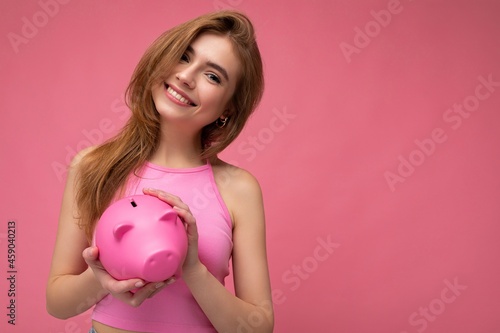 Portrait photo of happy positive smiling young beautiful attractive blonde woman with sincere emotions wearing pink summer top isolated over pink background with copy space and holding pink piggy box