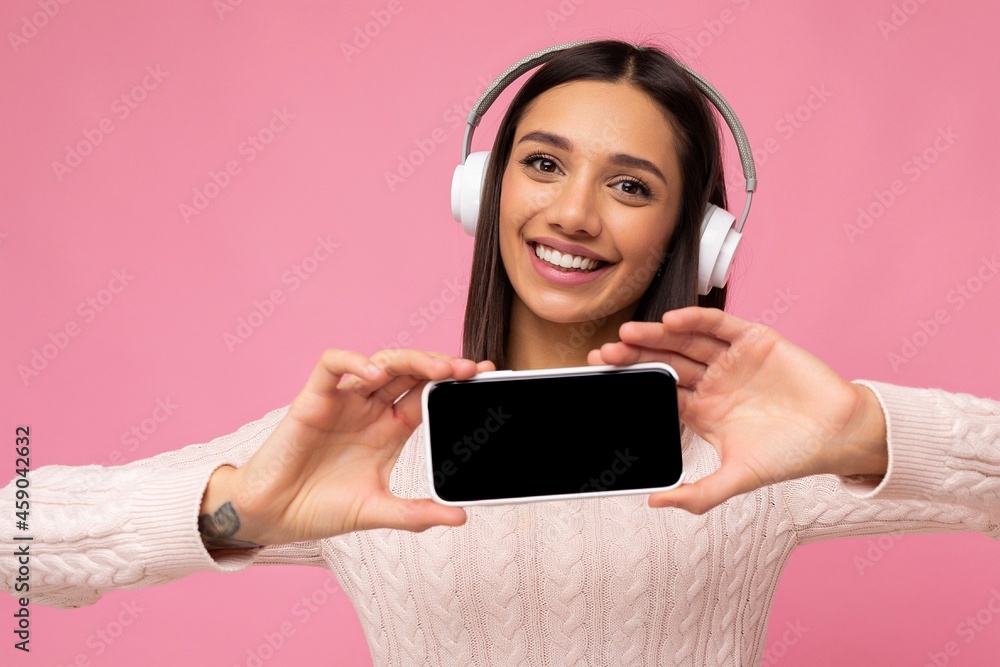 Closeup photo of beautiful happy smiling young woman wearing stylish casual outfit isolated on background wall holding and showing mobile phone with empty display for mockup wearing white bluetooth Closeup photo of beautiful happy smiling young woman wearing stylish casual outfit isolated on background wall holding and showing mobile phone with empty display for mockup wearing white bluetooth