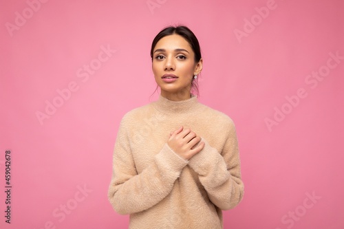 Portrait of young positive beautiful brunette woman with sincere emotions wearing beige pullover isolated over pink background with copy space and holding hand on chest