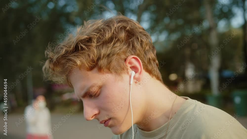 Young blond cool boy with earrings sitting on the beach and listening to music with headphones outside.