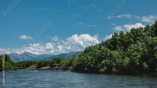 An inflatable boat with people is floating on the blue river. Lush green vegetation on the banks. A mountain range against the background of an azure sky and clouds. A sunny summer day. Kamchatka.