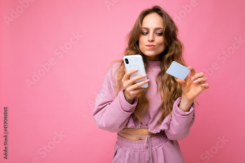 Beautiful sexy positive young blonde curly woman wearing pink clothes isolated over pink background using mobile phone paying online shopping through credit card looking at smartphone display