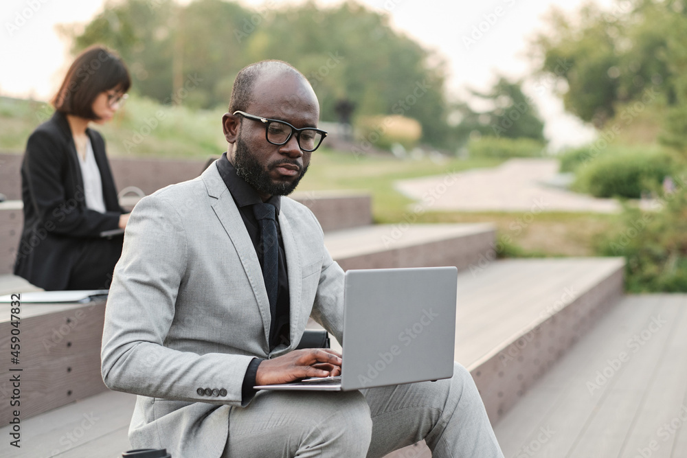 Horizontal medium portrait of serious mature African American man with beard on face wearing elegant suit sitting in urban park using laptop Horizontal medium portrait of serious mature African American man with beard on face wearing elegant suit sitting in urban park using laptop