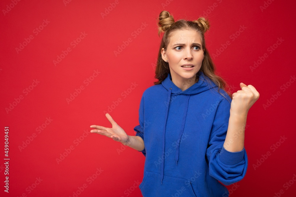Photo of emotional agressive young beautiful blonde woman with two horns with sincere emotions wearing hipster bright blue hoodie isolated over red background with free space and showing fist Photo of emotional agressive young beautiful blonde woman with two horns with sincere emotions wearing hipster bright blue hoodie isolated over red background with free space and showing fist