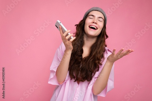 Positive attractive young brunette woman wearing stylish pink shirt and grey hat isolated over pink background holding in hand and using mobile phone