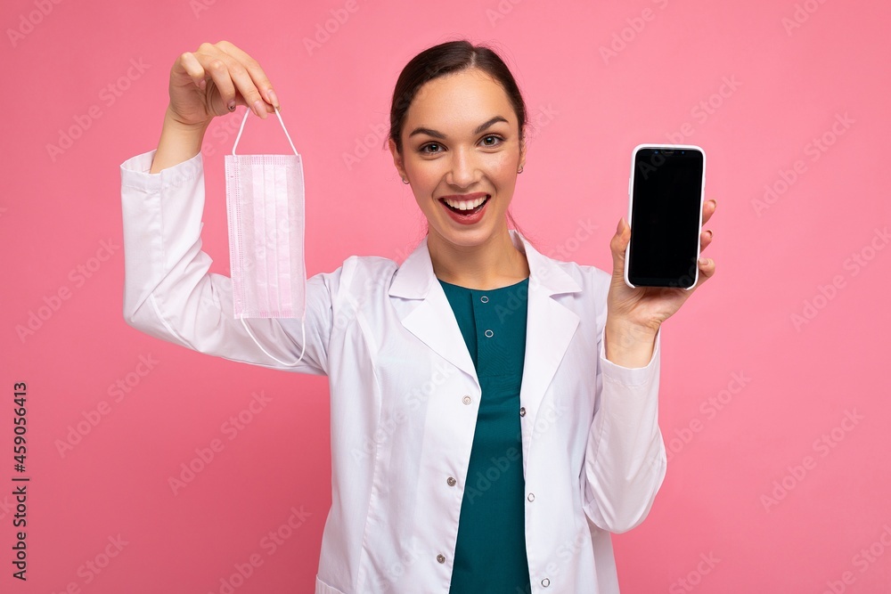 Photo of smiling medical adult female person wearing white coat holding mask showing modern mobile phone isolated over pink backgroung looking at camera Photo of smiling medical adult female person wearing white coat holding mask showing modern mobile phone isolated over pink backgroung looking at camera