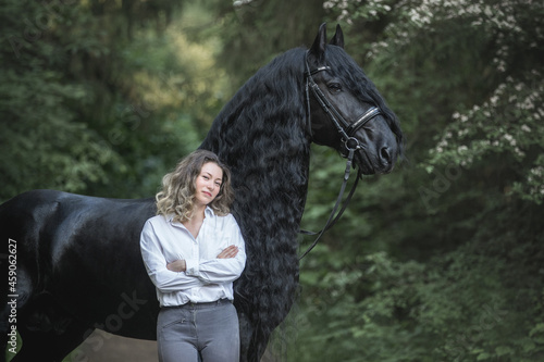 Beautiful young lady posing with a purebred friasian horse in the forest.