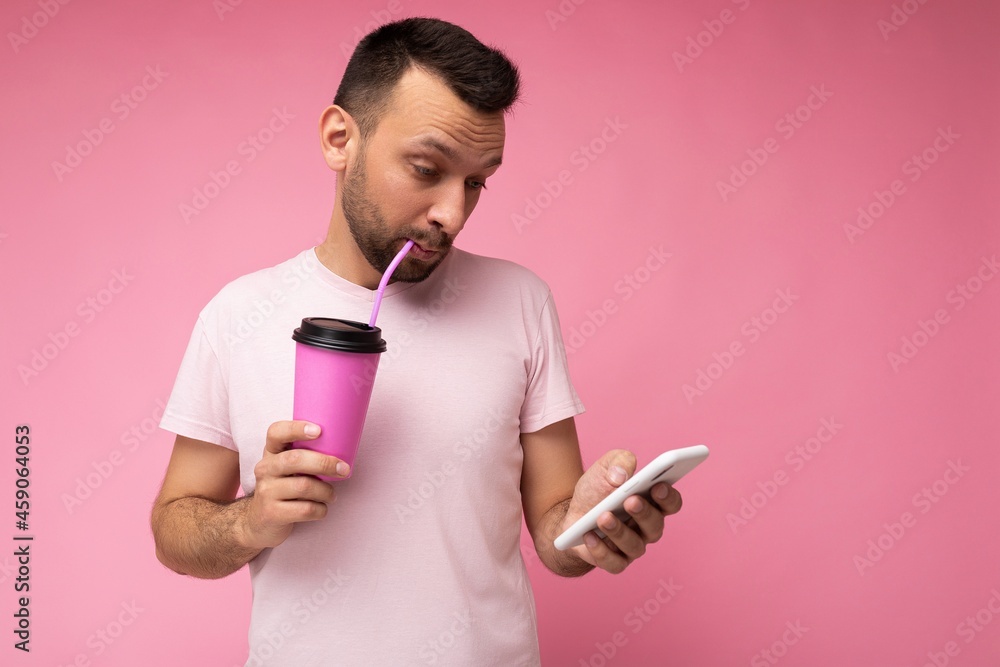 Photo shot of handsome positive good looking young man wearing casual stylish outfit poising isolated on background with empty space holding in hand and using mobile phone messaging sms looking at Photo shot of handsome positive good looking young man wearing casual stylish outfit poising isolated on background with empty space holding in hand and using mobile phone messaging sms looking at