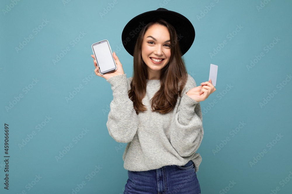 Beautiful positive smiling young brunette woman wearing black hat and grey sweater isolated over blue background holding credit card and mobile phone with empty display for mockup looking at camera Beautiful positive smiling young brunette woman wearing black hat and grey sweater isolated over blue background holding credit card and mobile phone with empty display for mockup looking at camera