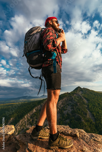 A male traveler in a red hat with a backpack is talking on the phone against the background of mountains. A man in hiking clothes talks on a smartphone at sunset in the mountains. Phone conversation