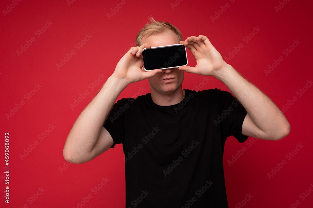 Portrait of handsome blonde young man wearing black t-shirt standing isolated on red background holding mobile phone showing smartphone in hand with empty display for cutout and mockup Portrait of handsome blonde young man wearing black t-shirt standing isolated on red background holding mobile phone showing smartphone in hand with empty display for cutout and mockup