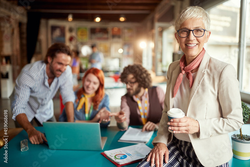 An elderly female boss in the office is posing for a photo while working with her young colleagues