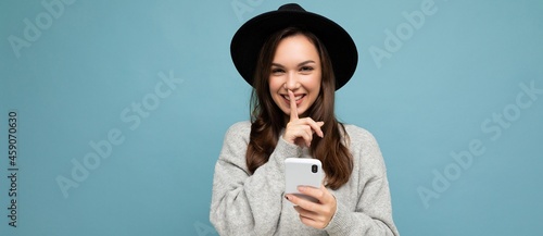 Panoramic photo of Attractive young smiling woman wearing black hat and grey sweater holding smartphone looking at camera showing shhh gesture isolated on background photo
