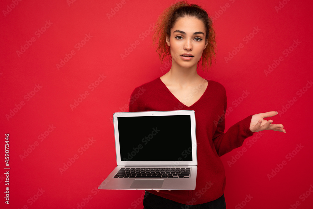 Beautiful charming fascinating young curly dark blond woman wearing red sweater standing isolated over red wall background holding computer laptop with empty copy space mock up pointing to the side Beautiful charming fascinating young curly dark blond woman wearing red sweater standing isolated over red wall background holding computer laptop with empty copy space mock up pointing to the side