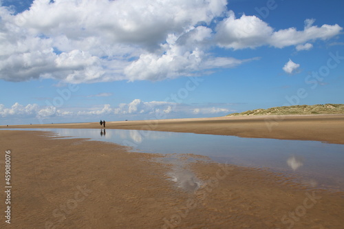 Beautiful landscape of Holkham Norfolk uk sandy beach with couple on holiday walk by ocean shore with white puffy clouds reflected in sea water lakes formed on the sand in low tide Summer blue skies © Carmina