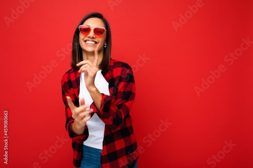 Photo of young beautiful happy smiling brunette woman wearing trendy white t-shirt and red check shirt and red sunglasses. Sexy carefree female person posing isolated near red wall in studio with free