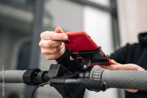 A woman's hand touches the screen of a smartphone mounted on the steering wheel of an electric scooter using a holder. photo