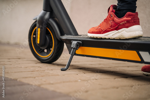 Female feet in red sneakers stand on the deck of the electric scooter, getting ready to start moving (focus on parking stand, foot)