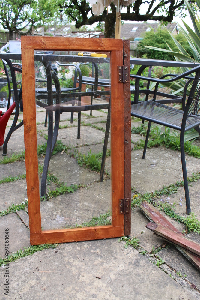 Close up of home made carpentry greenhouse window in glass and stained wood, the renovated oblong pine wooden frame stood patio outdoors garden with metal furniture background Close up of home made carpentry greenhouse window in glass and stained wood, the renovated oblong pine wooden frame stood patio outdoors garden with metal furniture background